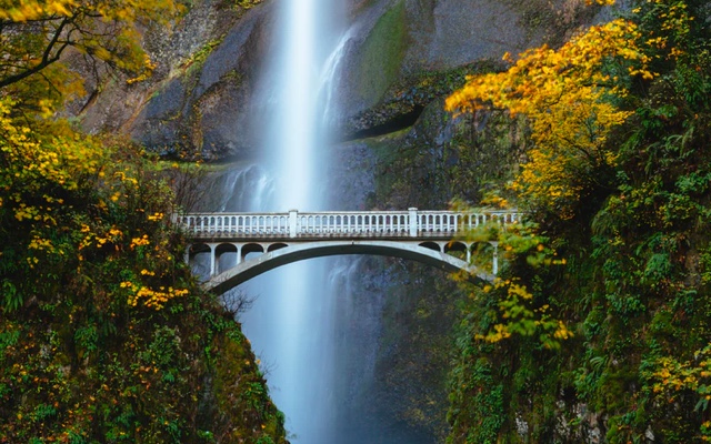 A side view of a bridge that arches over a ravine with a waterfall behind it